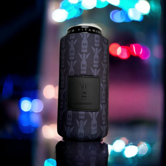 A beverage can wrapped in a dark gray bomber sleeve with a repeating bomber airplane pattern and a central black patch, set against a blurred night scene with colorful bokeh lights.