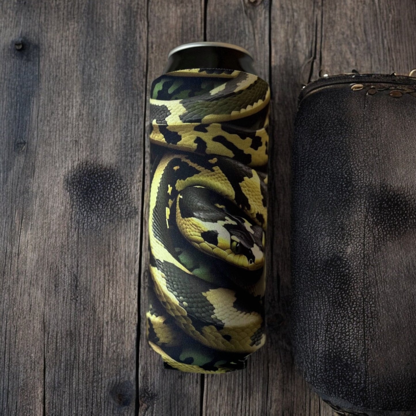 Bomber Venom AI Snake 4mm neoprene can insulator with yellow, green, and black viper scale pattern resting on a rustic dark wood surface.