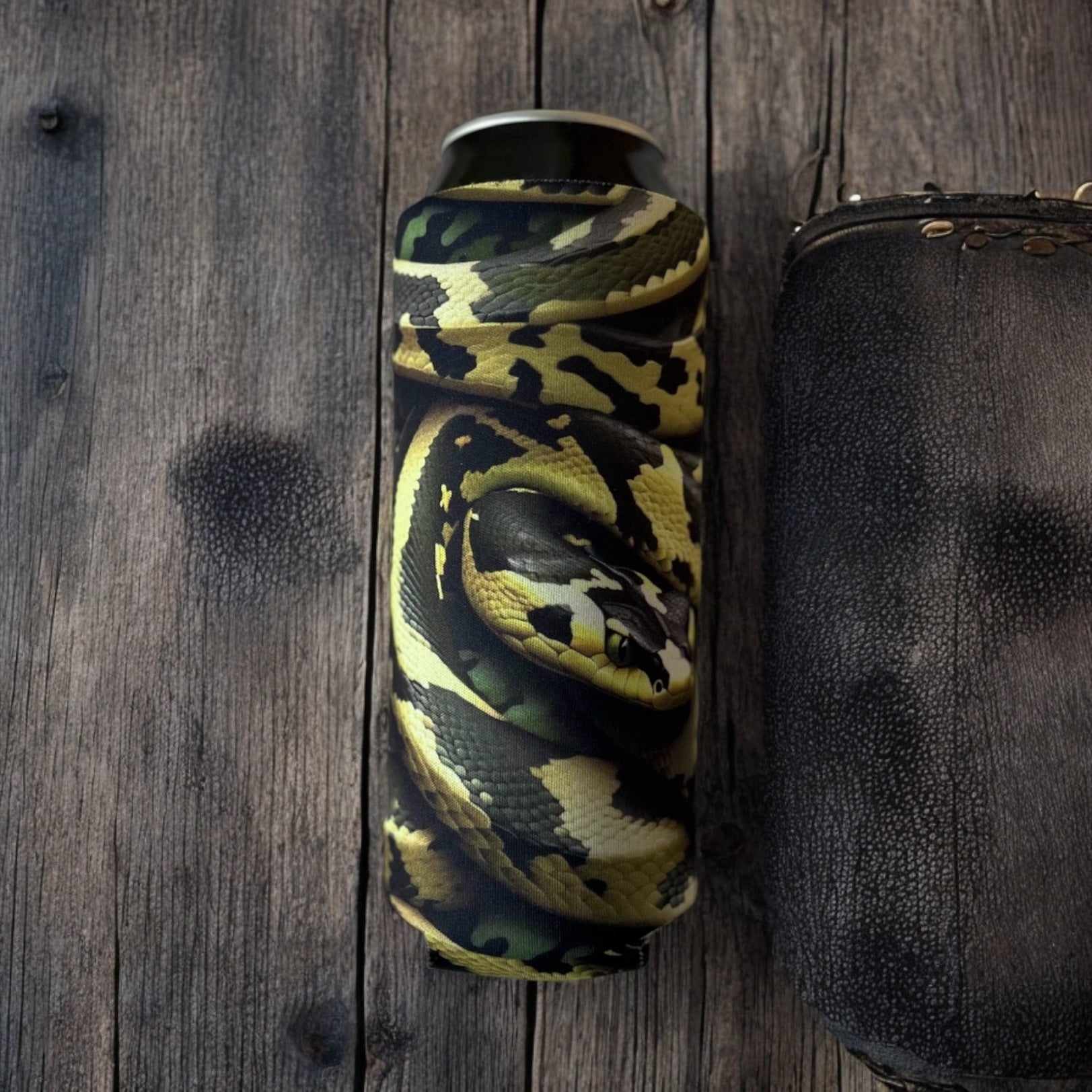 Bomber Venom AI Snake 4mm neoprene can insulator with yellow, green, and black viper scale pattern resting on a rustic dark wood surface.