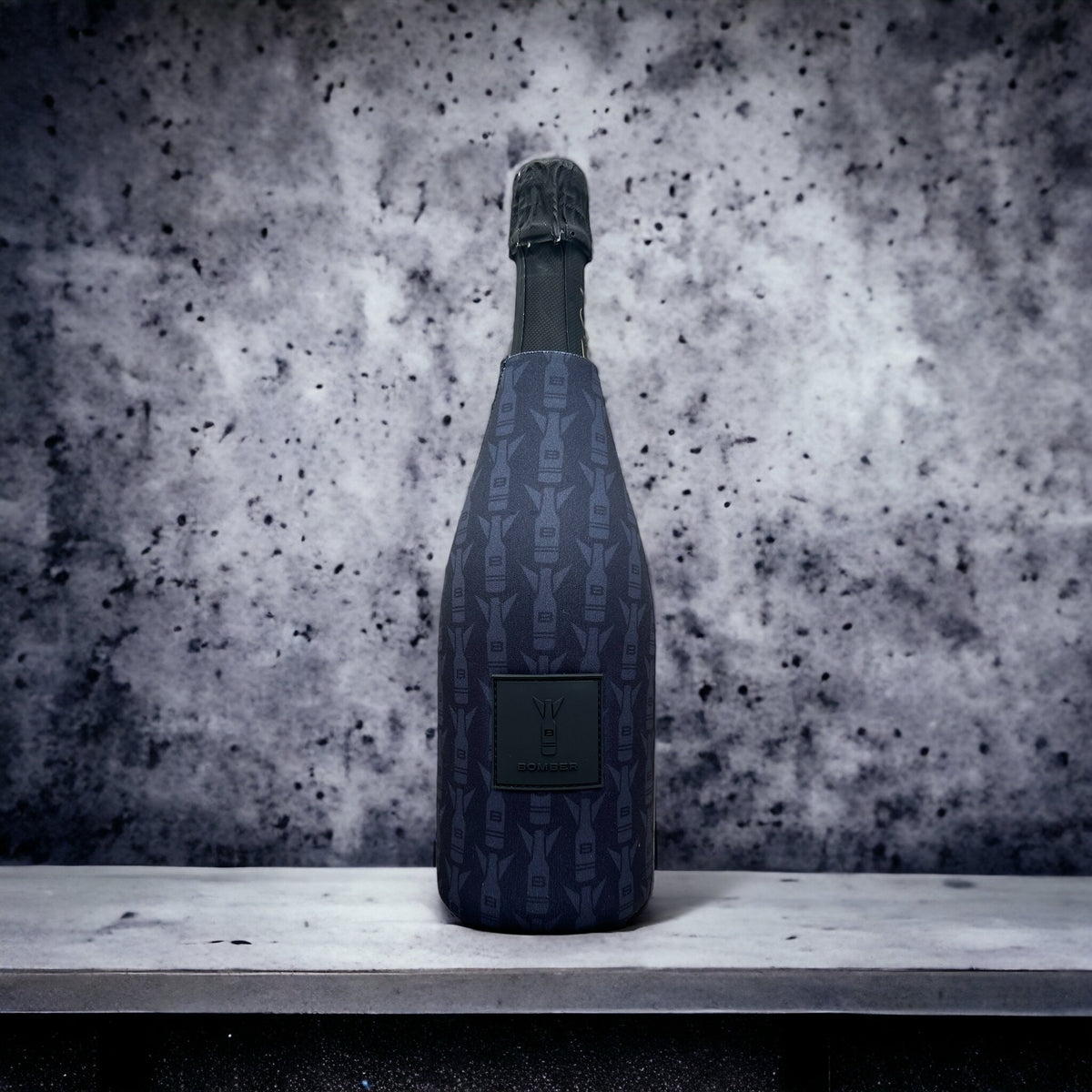 A dark blue patterned champagne bottle with a black foil top, standing on a light gray stone ledge against a textured gray and white speckled background.
