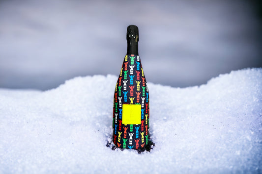 A champagne bottle standing upright in deep snow, wrapped in a black fabric cover printed with a colorful repeating pattern of stylized figures, with a blank yellow label on the front.