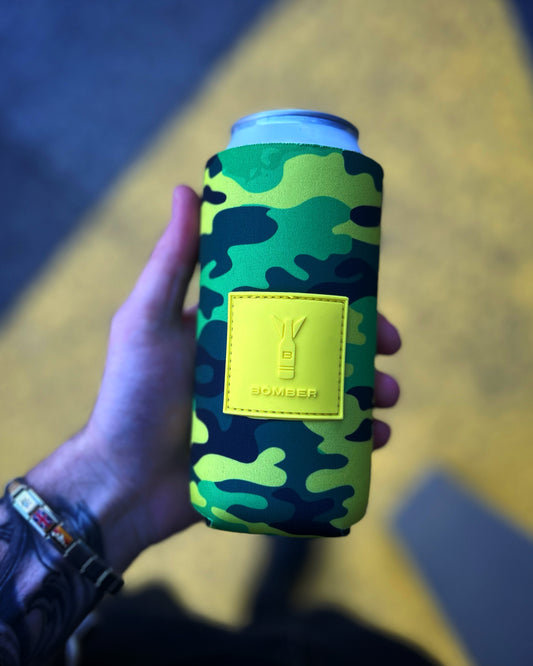 A hand holding a can cooler sleeve with a green and yellow camouflage pattern and a yellow 'BOMBER' logo patch, set against a blurred yellow and blue background.